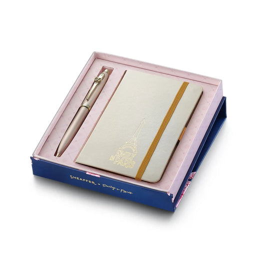 Sheaffer 100 Emily in Paris gift set featuring a champagne gold ballpoint pen with a bow-shaped clip emblem and a matching hardbound small notebook in a premium presentation box. Model G2937751-4