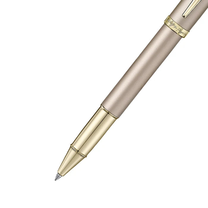 The forepart of a sleek Sheaffer 100  Emily in Paris rollerball pen in a metallic champagne gold lacquer finish with champagne gold  trims and adorned with a bow-shaped emblem on the clip. Model E1937751