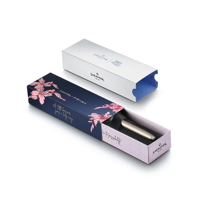 The box of a sleek Sheaffer 100  Emily in Paris rollerball pen in a metallic champagne gold lacquer finish with champagne gold  trims and adorned with a bow-shaped emblem on the clip. Model E1937751